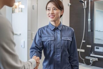 青い制服を着た人が笑顔で別の人と握手しています。彼らはモダンなバスルームに立っており、背景にはシャワーとさまざまな備品が見えます。このシーンはプロフェッショナルでフレンドリーな雰囲気を反映しており、中野区排水管修理のようなサービスを示唆しています。
